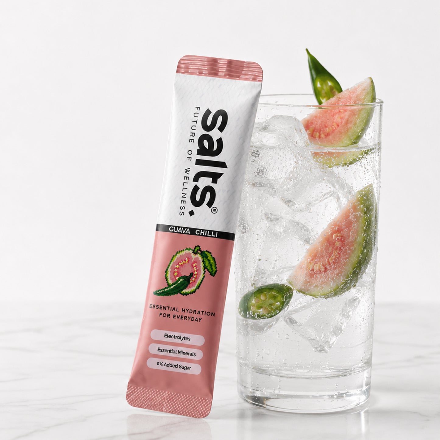 A single packet of Salts Guava Chilli electrolyte drink mix stands next to a tall glass of water with ice, watermelon slices, and a chili pepper on a light marble surface against a white background.